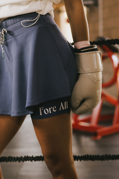 Person wearing an indigo skirt with 'Fore All' text and beige boxing gloves in a gym setting.