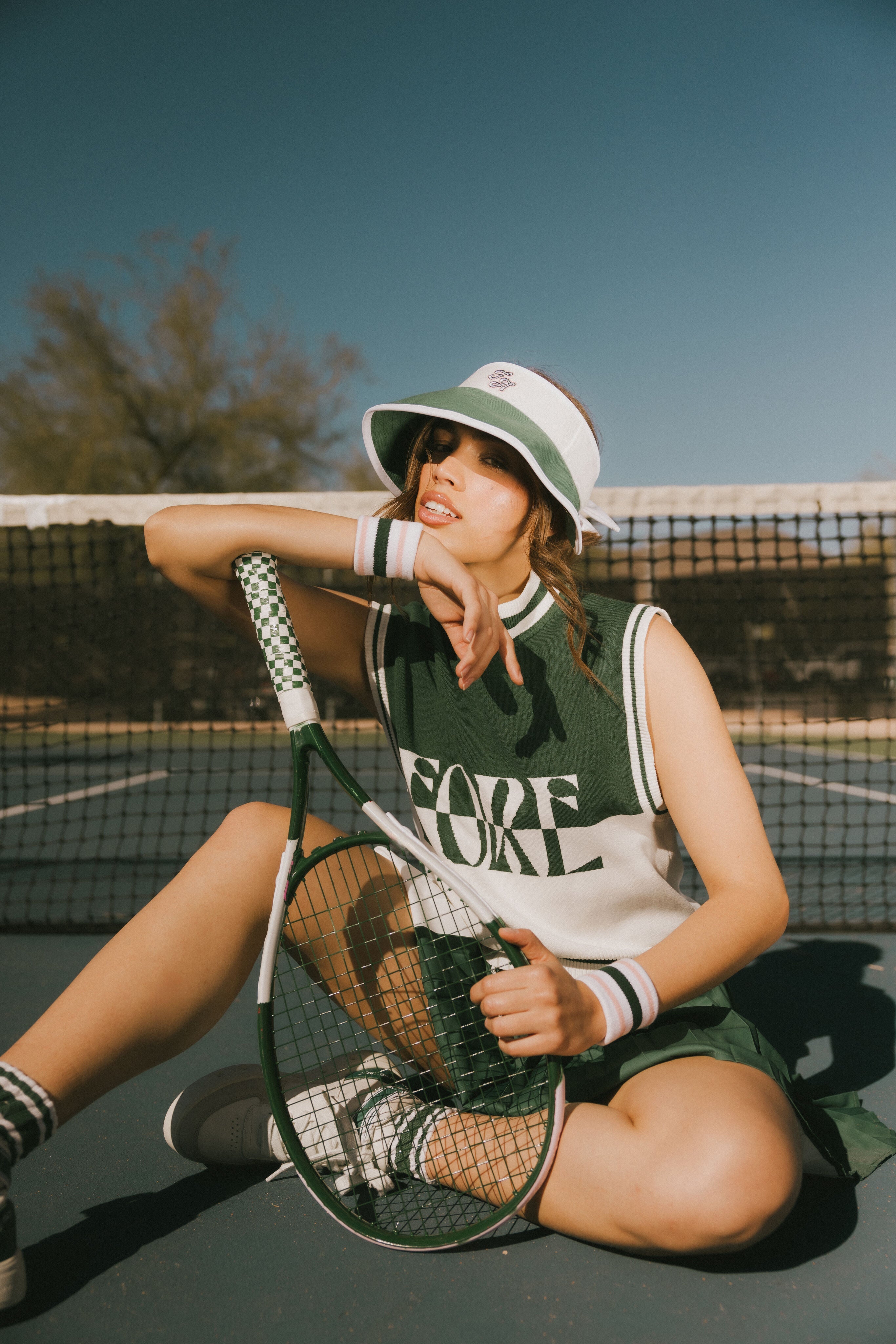Person sitting on a tennis court holding a racket, wearing a green and white fore sleeveless sweater.