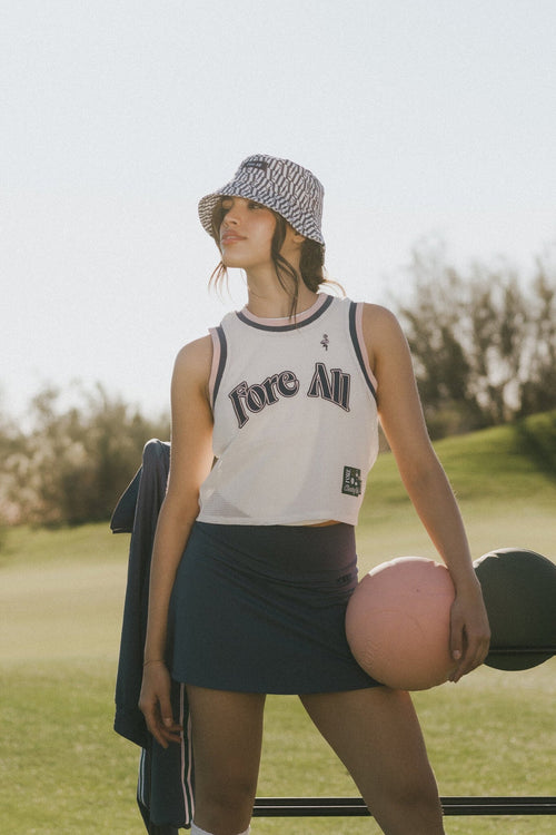 Woman in a white tank top with 'Fore All' text, indigo skirt, and patterned hat standing on a golf course.