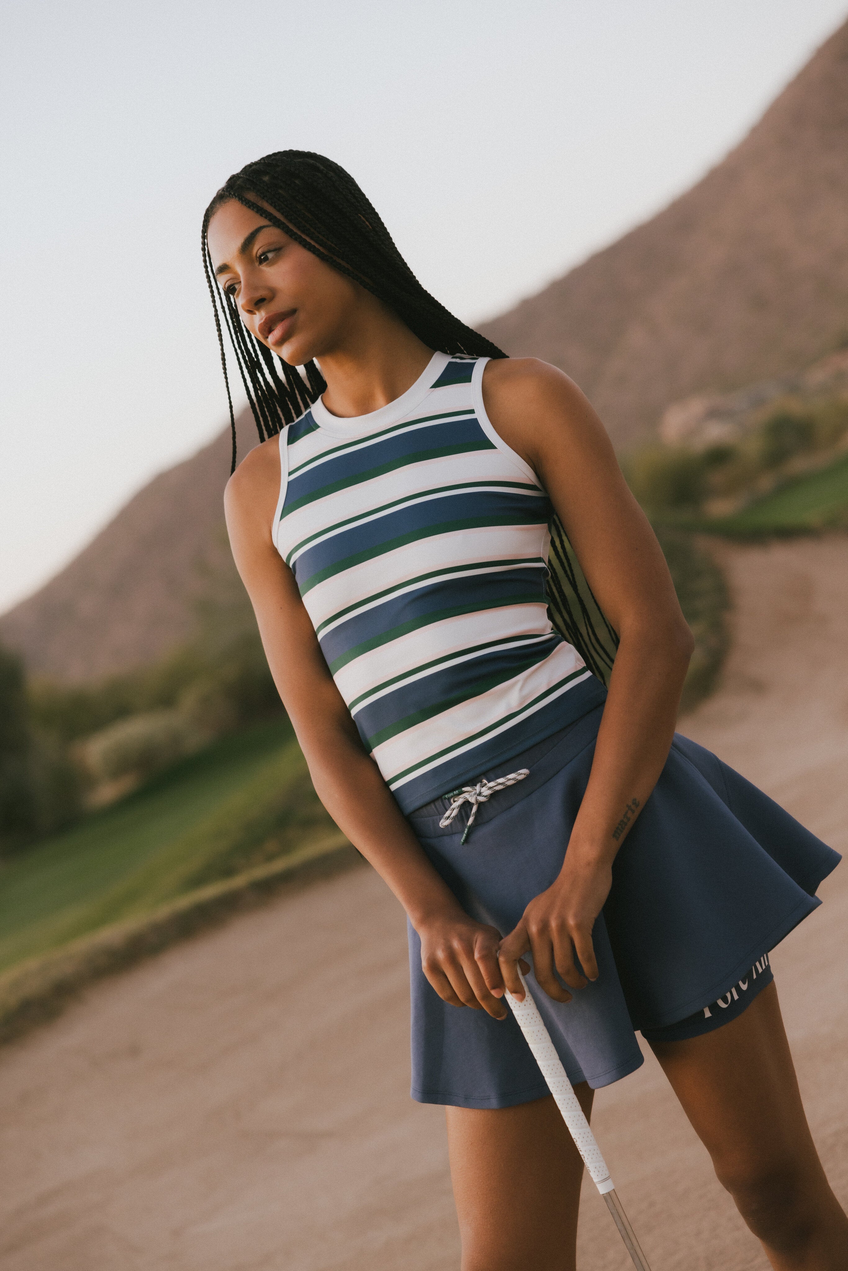Woman in a striped sleeveless top and blue skirt holding a golf club with a golf course in the background