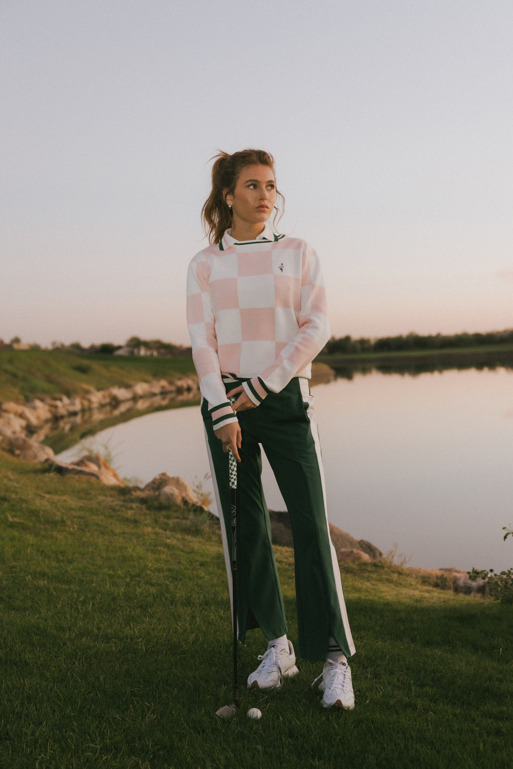 Woman in a checkered sweater and green pants holding a golf club on a golf course.