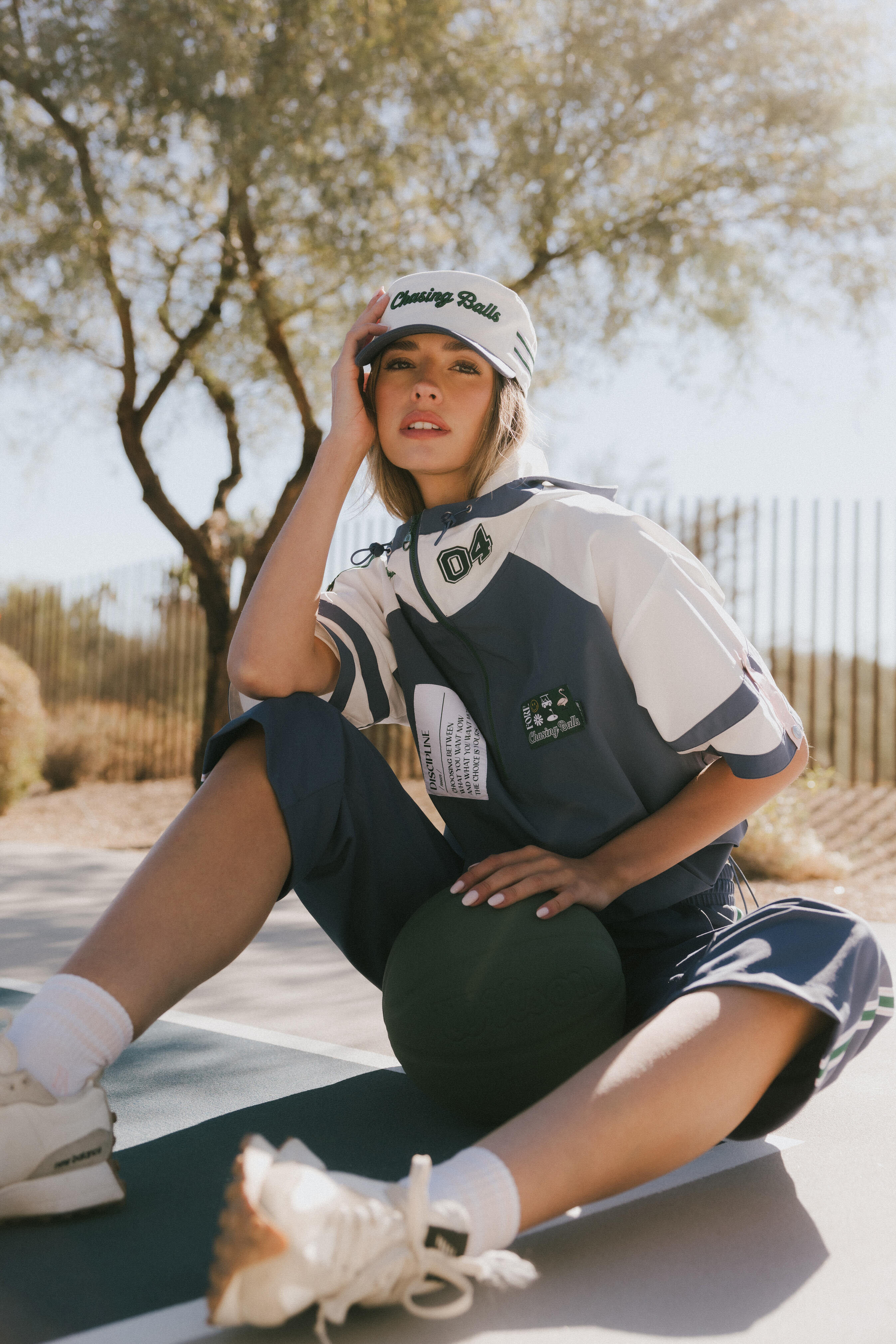 Woman sitting on a court wearing an indigo and white outfit with a cap, outdoors.