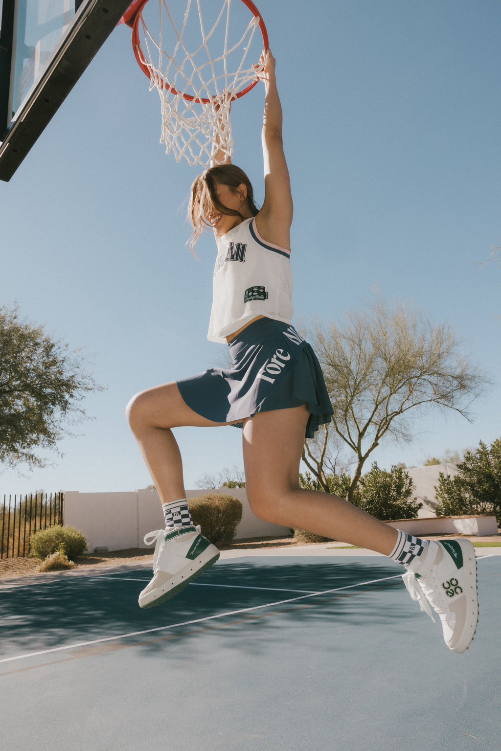 A woman wearing a white sleeveless tank top with 'Fore All' text and a flamingo logo on the chest, paired with an indigo skirt.