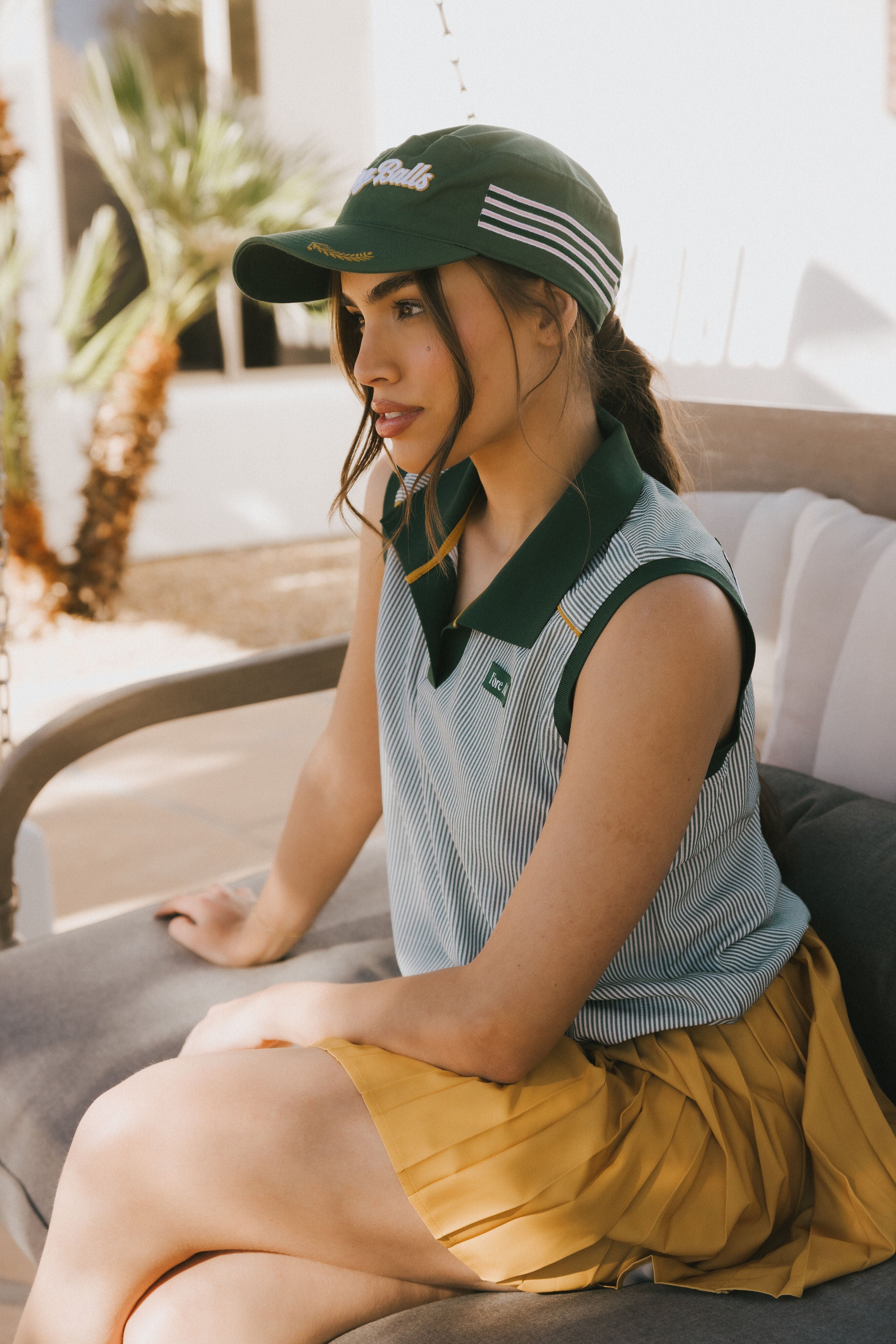 Woman wearing a green cap and sleeveless shirt sitting outdoors with plants in the background