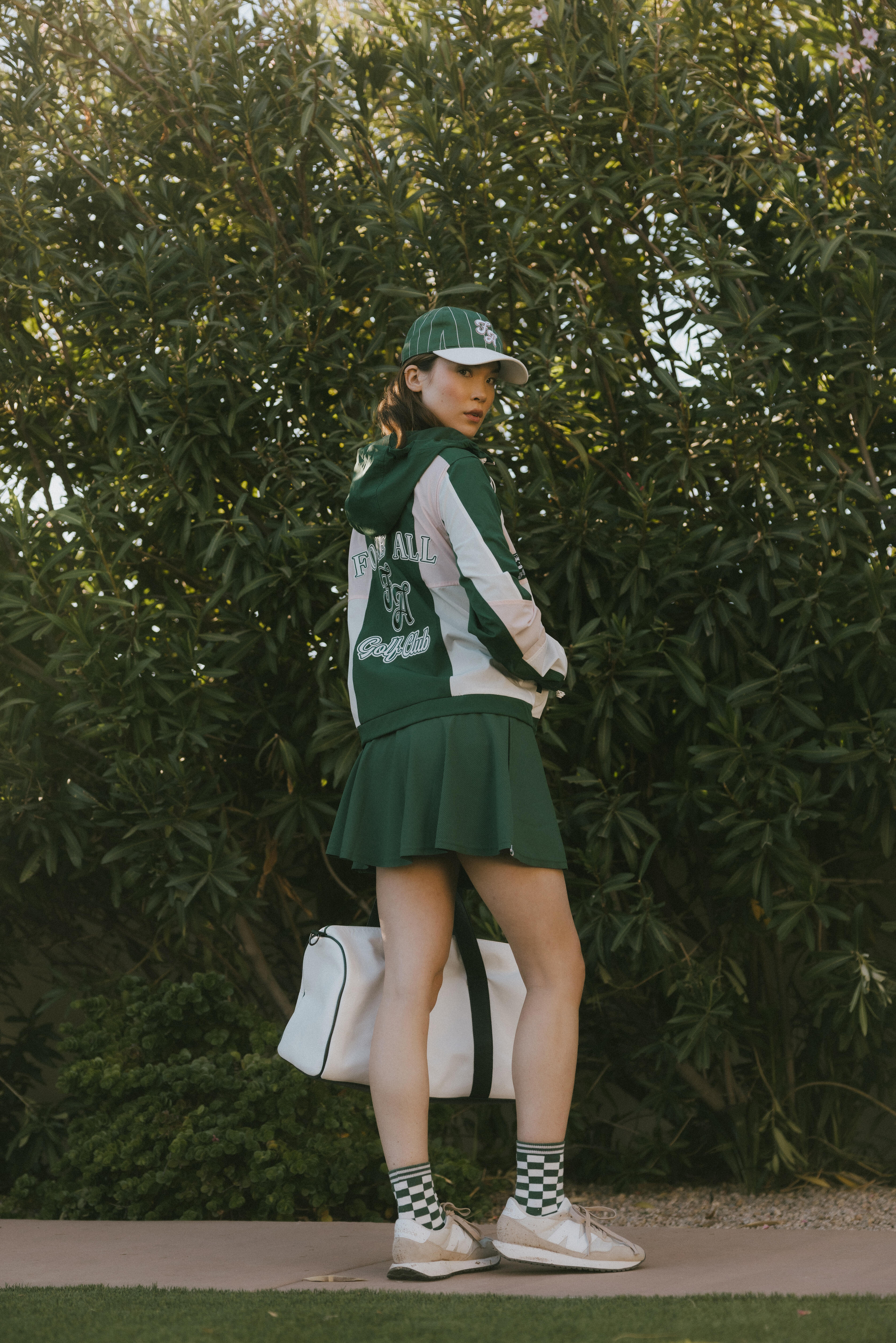 A woman wearing a long-sleeved green and pink warm-up jacket with a hood, zipper pockets, and colorblocking. She also sports a matching green cap.