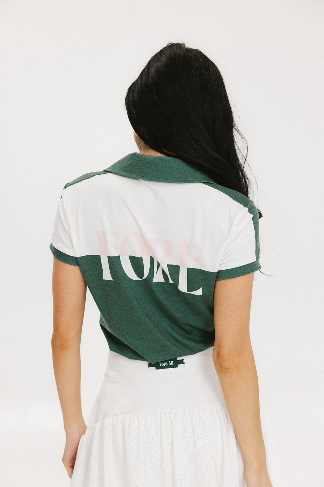 A woman wearing a color block green polo shirt with white front and back yoke detailing, paired with a white skirt.