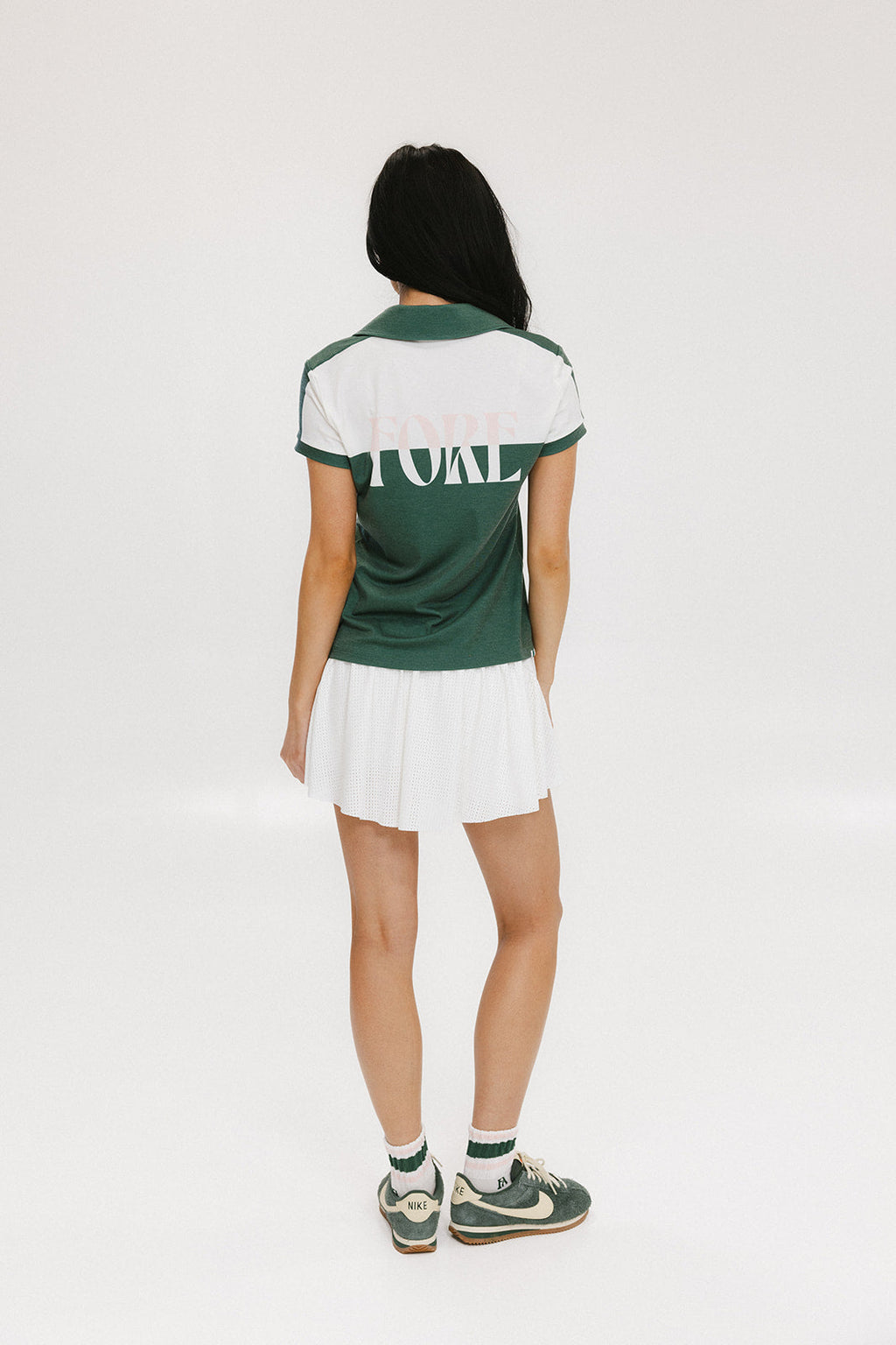 A woman wearing a color block green polo shirt with white front and back yoke detailing, paired with a white skirt.