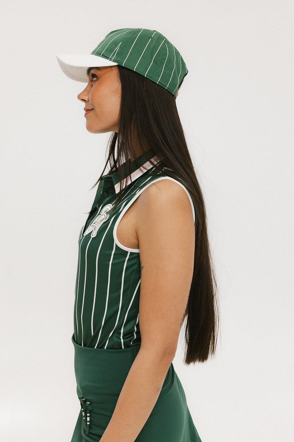 A female model wearing a sleeveless green pinstripe polo top with an applique logo on the chest and a matching green skirt, paired with a green cap.