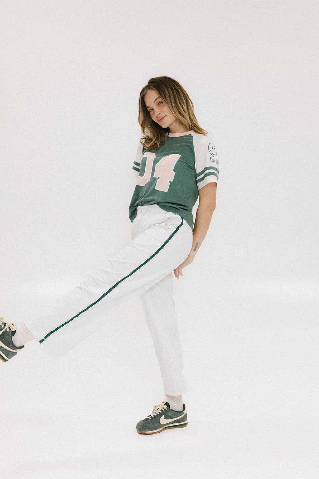 A woman posing with one leg lifted, wearing a green and white t-shirt with the text 'CHASING BALLS' on the back, paired with white pants and green sneakers.