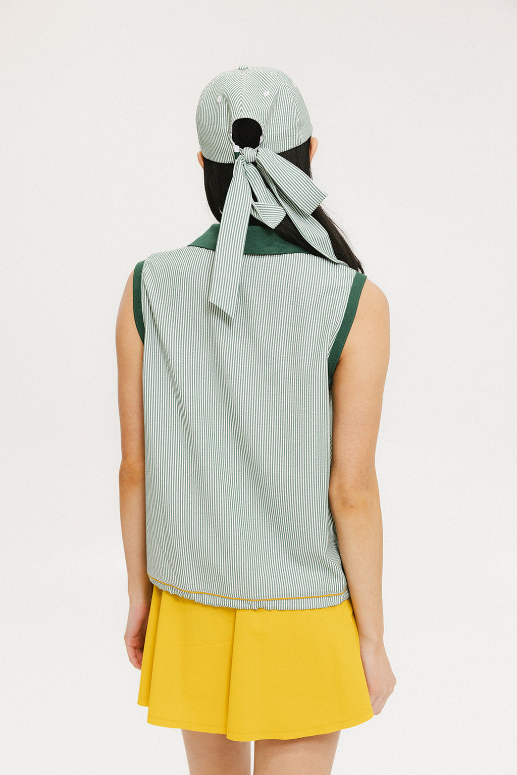 A woman wearing a green striped seersucker polo top with a collar, sleeveless with front yoke and piping accents, paired with a yellow skirt and a matching green cap.