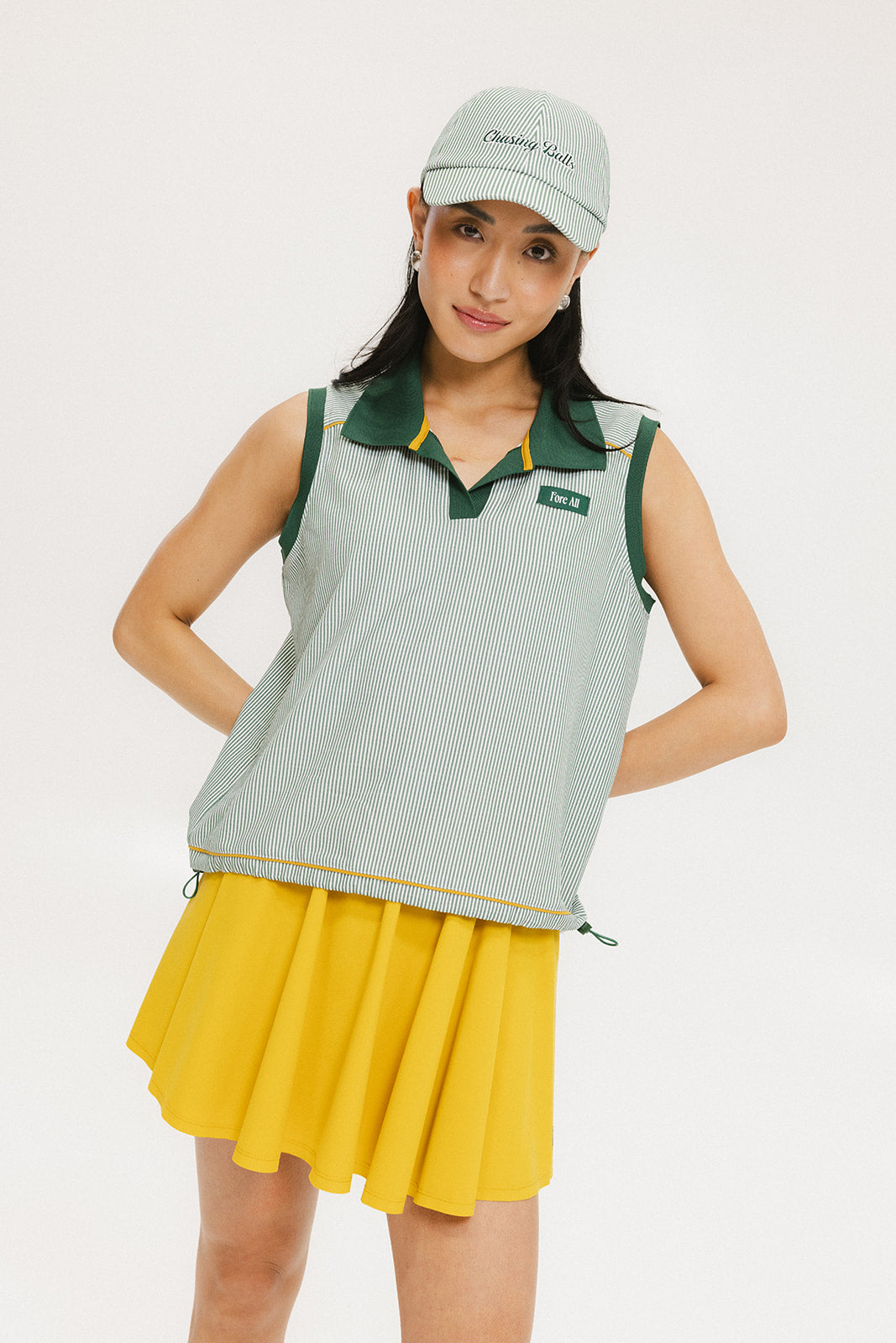 A woman wearing a green striped seersucker polo top with a collar, sleeveless with front yoke and piping accents, paired with a yellow skirt and a matching green cap.