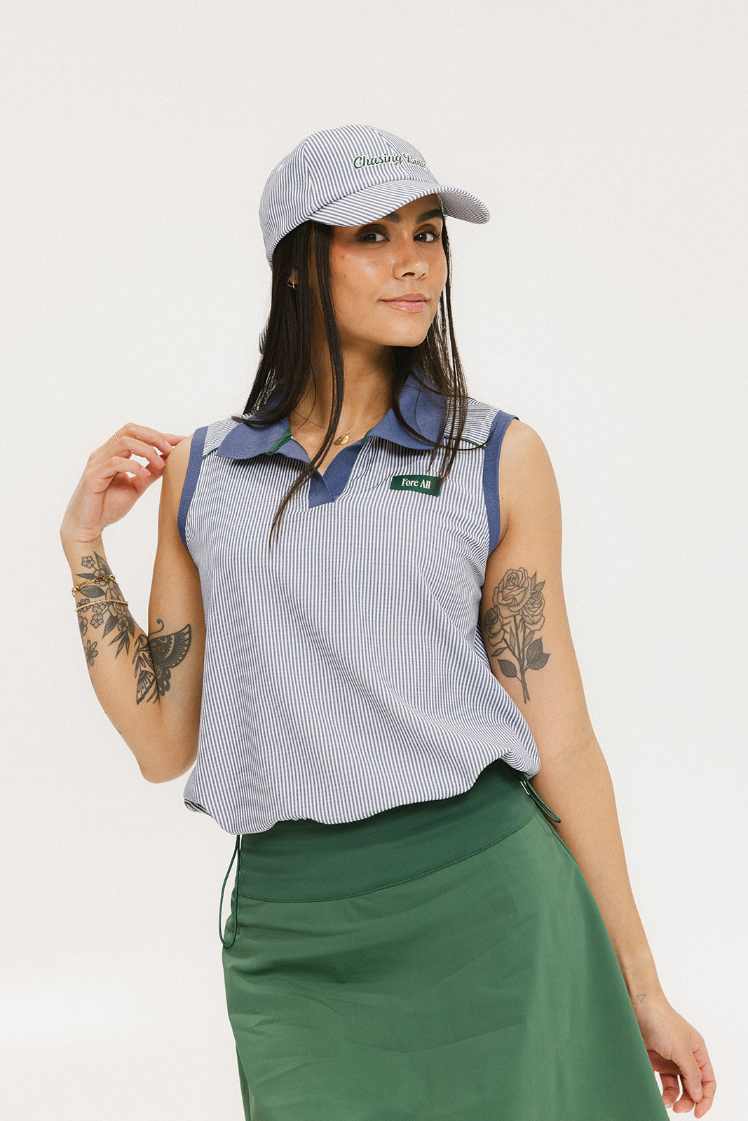 A woman wearing a sleeveless indigo-striped seersucker polo shirt with collar, sleeve yoke, and piping accents. The shirt has elastic toggles at the hem and side seam, and sleeveless appliqué logos.