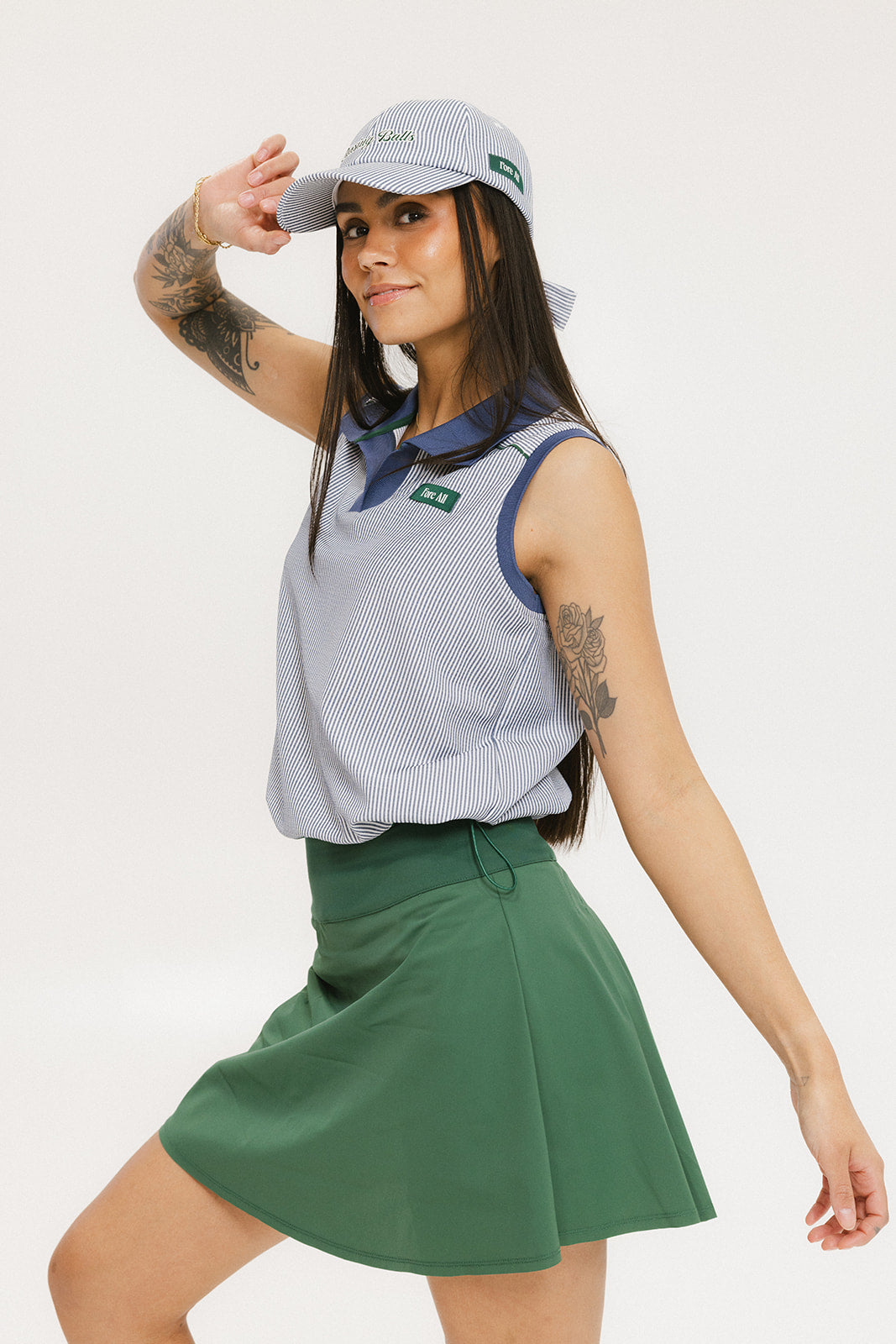 A woman wearing a sleeveless indigo-striped seersucker polo shirt with collar, sleeve yoke, and piping accents. The shirt has elastic toggles at the hem and side seam, and sleeveless appliqué logos.