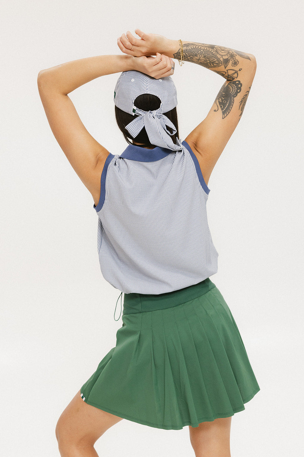 A woman wearing a sleeveless indigo-striped seersucker polo shirt with collar, sleeve yoke, and piping accents. The shirt has elastic toggles at the hem and side seam, and sleeveless appliqué logos.