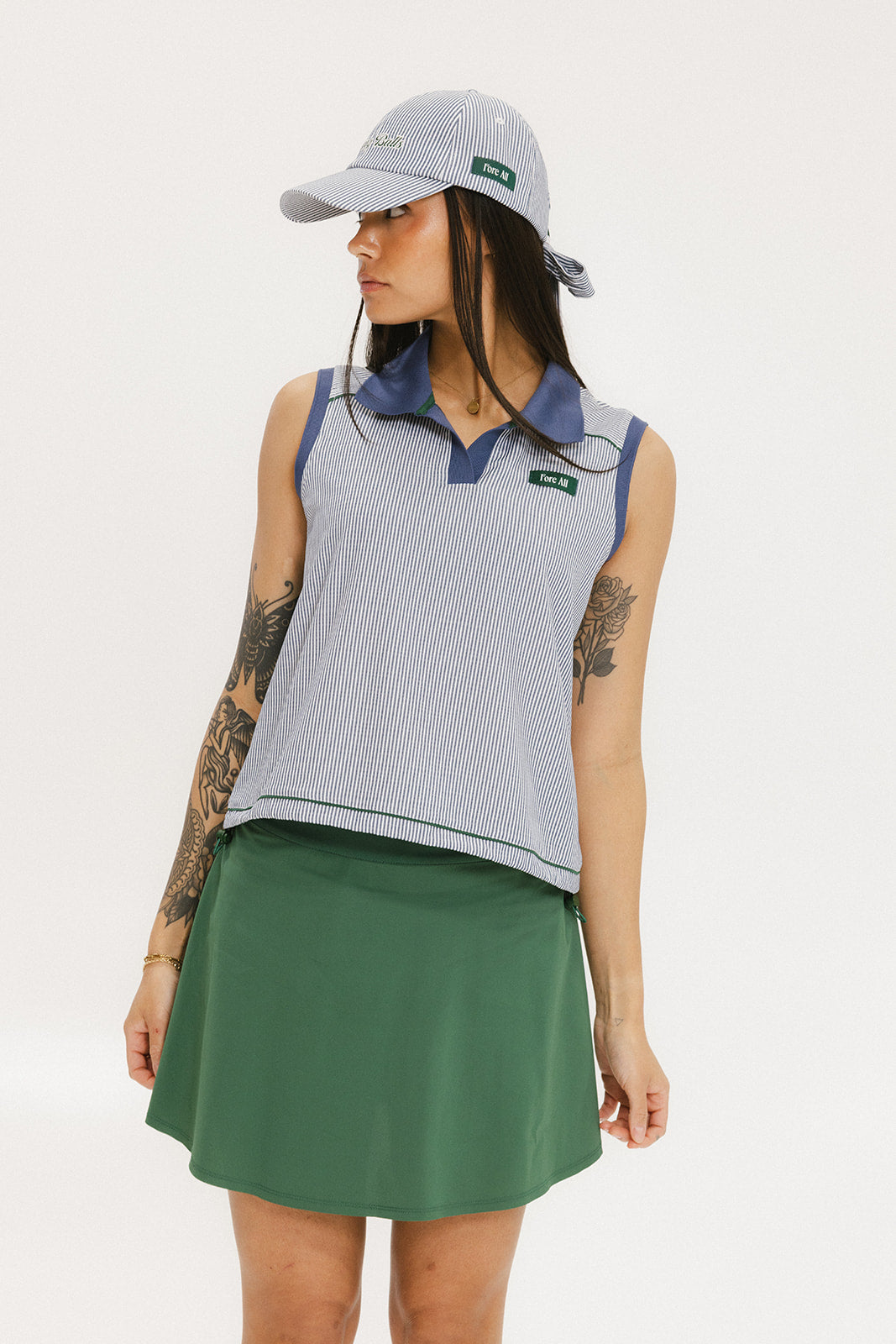 A woman wearing a sleeveless indigo-striped seersucker polo shirt with collar, sleeve yoke, and piping accents. The shirt has elastic toggles at the hem and side seam, and sleeveless appliqué logos.