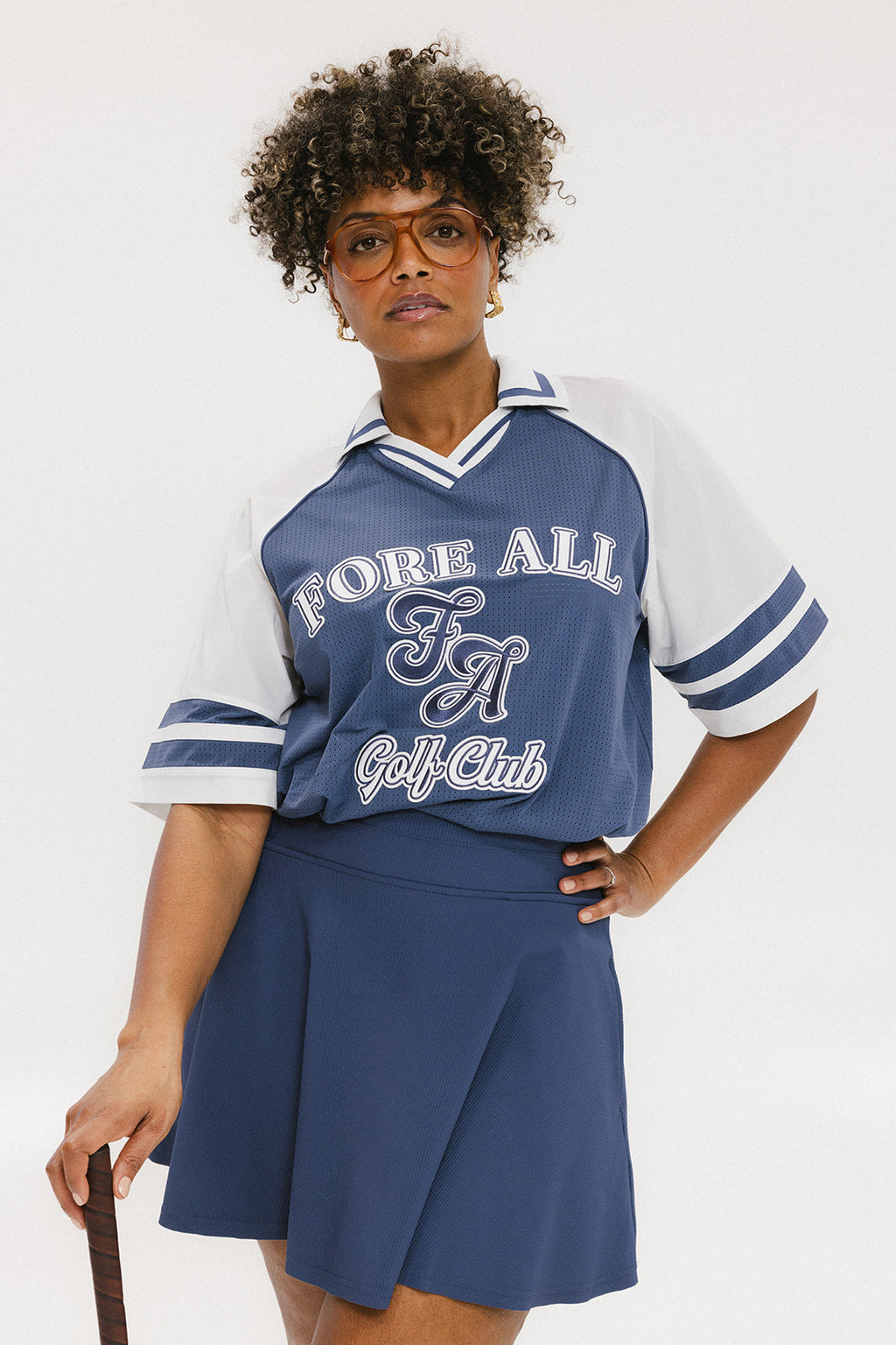 A person wearing a blue and white rugby-inspired polo shirt with Mesh body appliqué logos, a ribbed collar and placket, and elastic toggles at the side seam hem. The shirt has 'FORE ALL' text and a golf club logo on the front.