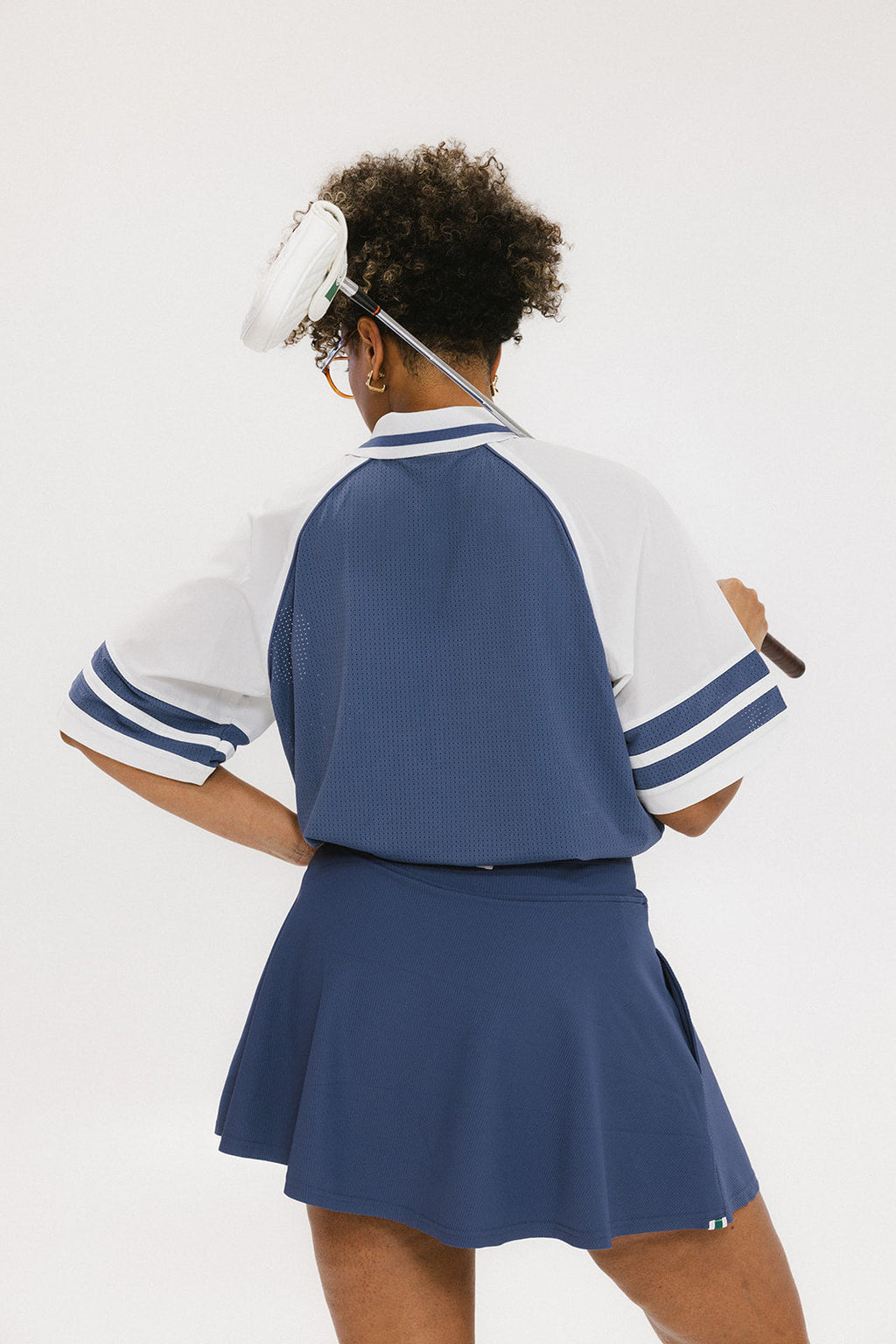 A person wearing a blue and white rugby-inspired polo shirt with Mesh body appliqué logos, a ribbed collar and placket, and elastic toggles at the side seam hem. The shirt has 'FORE ALL' text and a golf club logo on the front.