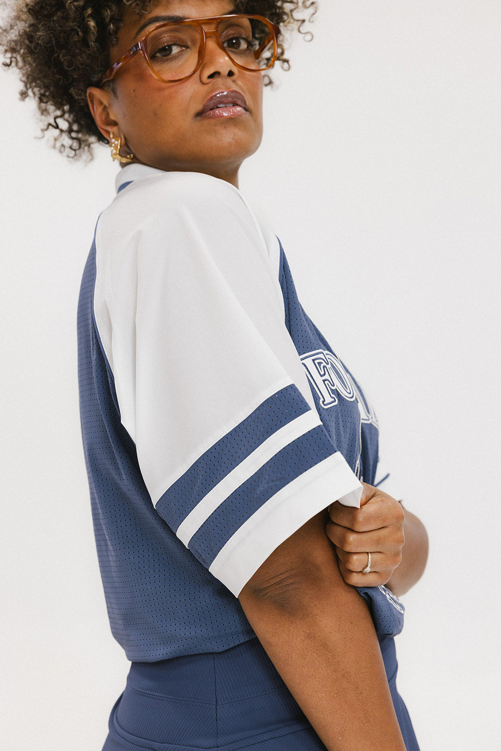 A person wearing a blue and white rugby-inspired polo shirt with Mesh body appliqué logos, a ribbed collar and placket, and elastic toggles at the side seam hem. The shirt has 'FORE ALL' text and a golf club logo on the front.