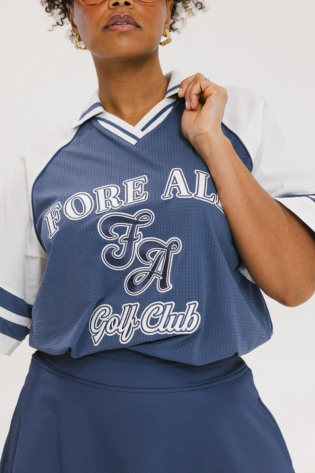A person wearing a blue and white rugby-inspired polo shirt with Mesh body appliqué logos, a ribbed collar and placket, and elastic toggles at the side seam hem. The shirt has 'FORE ALL' text and a golf club logo on the front.