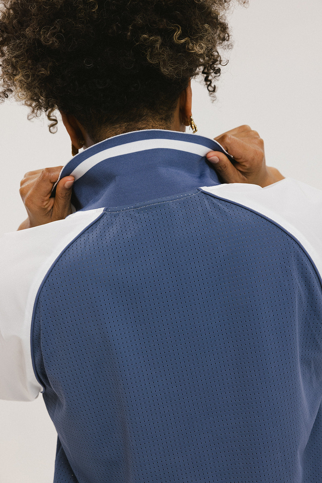 A person wearing a blue and white rugby-inspired polo shirt with Mesh body appliqué logos, a ribbed collar and placket, and elastic toggles at the side seam hem. The shirt has 'FORE ALL' text and a golf club logo on the front.