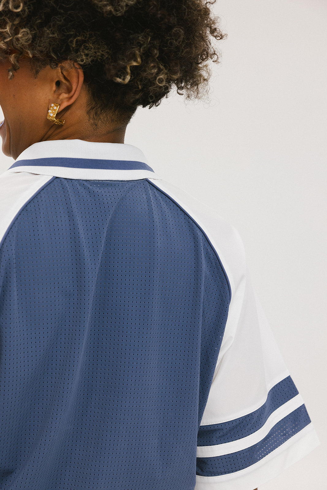 A person wearing a blue and white rugby-inspired polo shirt with Mesh body appliqué logos, a ribbed collar and placket, and elastic toggles at the side seam hem. The shirt has 'FORE ALL' text and a golf club logo on the front.