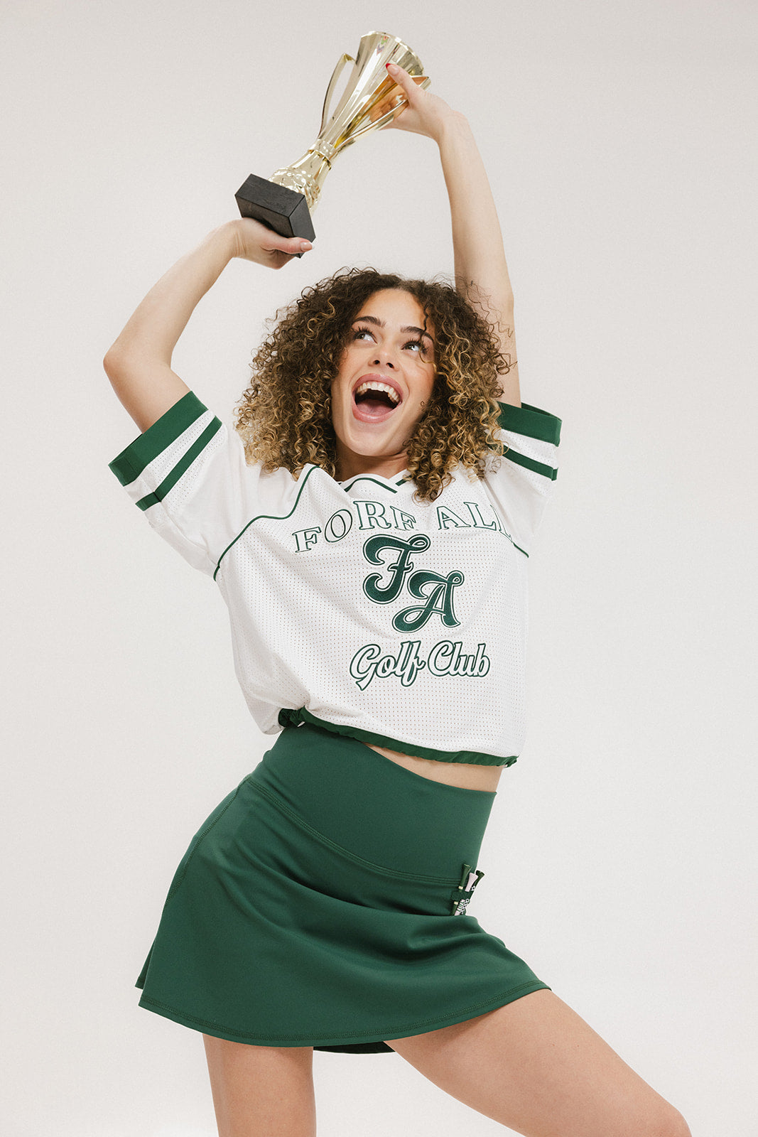 A woman wearing a white rugby-inspired polo shirt with green details and text that reads 'FORE ALL Golf Club'.