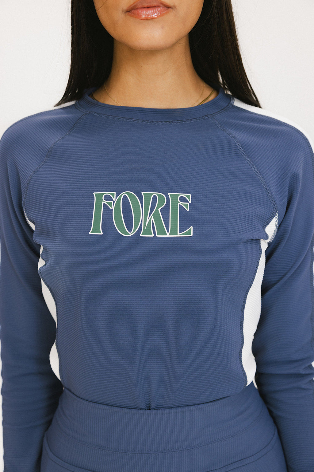 A female model wearing a long sleeve activewear top in blue and white with the text 'FORE' on the front, paired with a matching skirt.