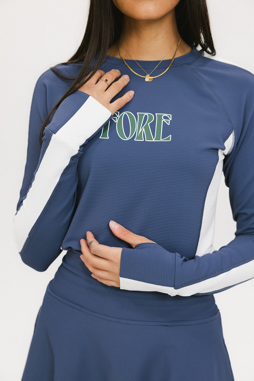 A female model wearing a long sleeve activewear top in blue and white with the text 'FORE' on the front, paired with a matching skirt and a visor with the text 'FORE'.