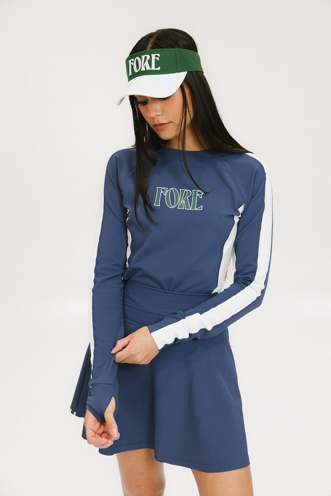 A female model wearing a long sleeve activewear top in blue and white with the text 'FORE' on the front, paired with a matching skirt and a visor with the text 'FORE'.