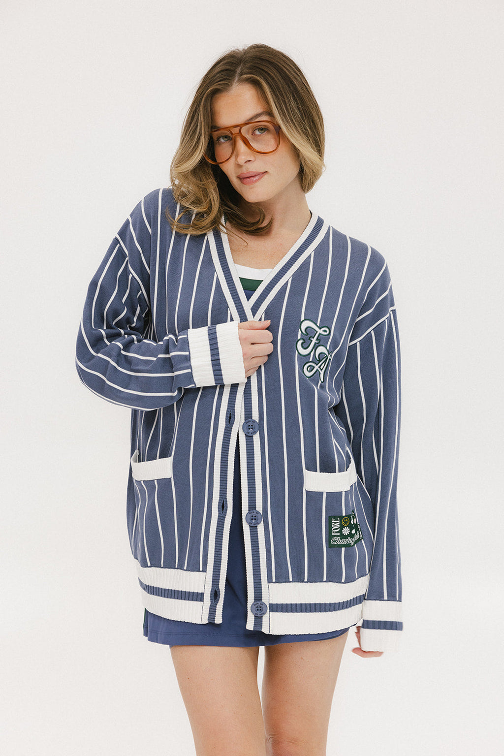 A woman wearing an oversized blue and white pinstripe cardigan with intarsia knit details and applique logos, paired with an indigo dress.