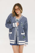 A woman wearing an oversized blue and white pinstripe cardigan with intarsia knit details and applique logos, paired with an indigo dress.