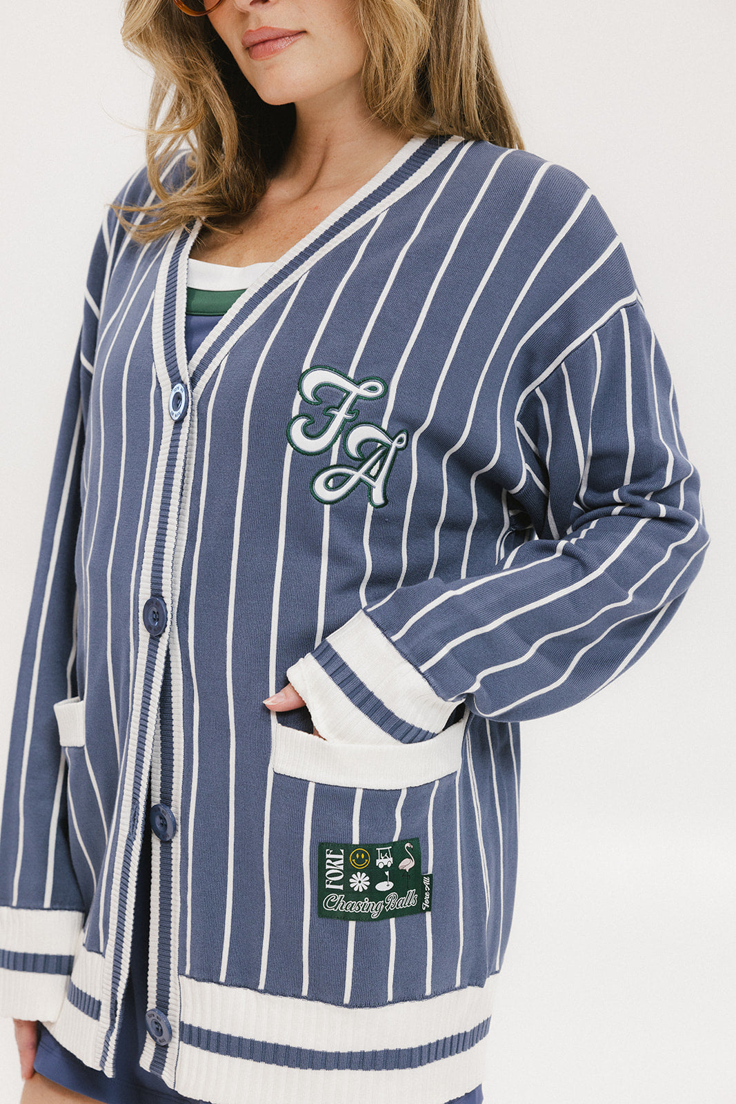 A woman wearing an oversized blue and white pinstripe cardigan with intarsia knit details and applique logos, paired with an indigo dress.