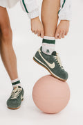 A person standing on one leg placing a retro ankle sock into their shoe, with the sock displaying retro-inspired stripes in pink and green colors, and a logo 'FA' on the cuff.