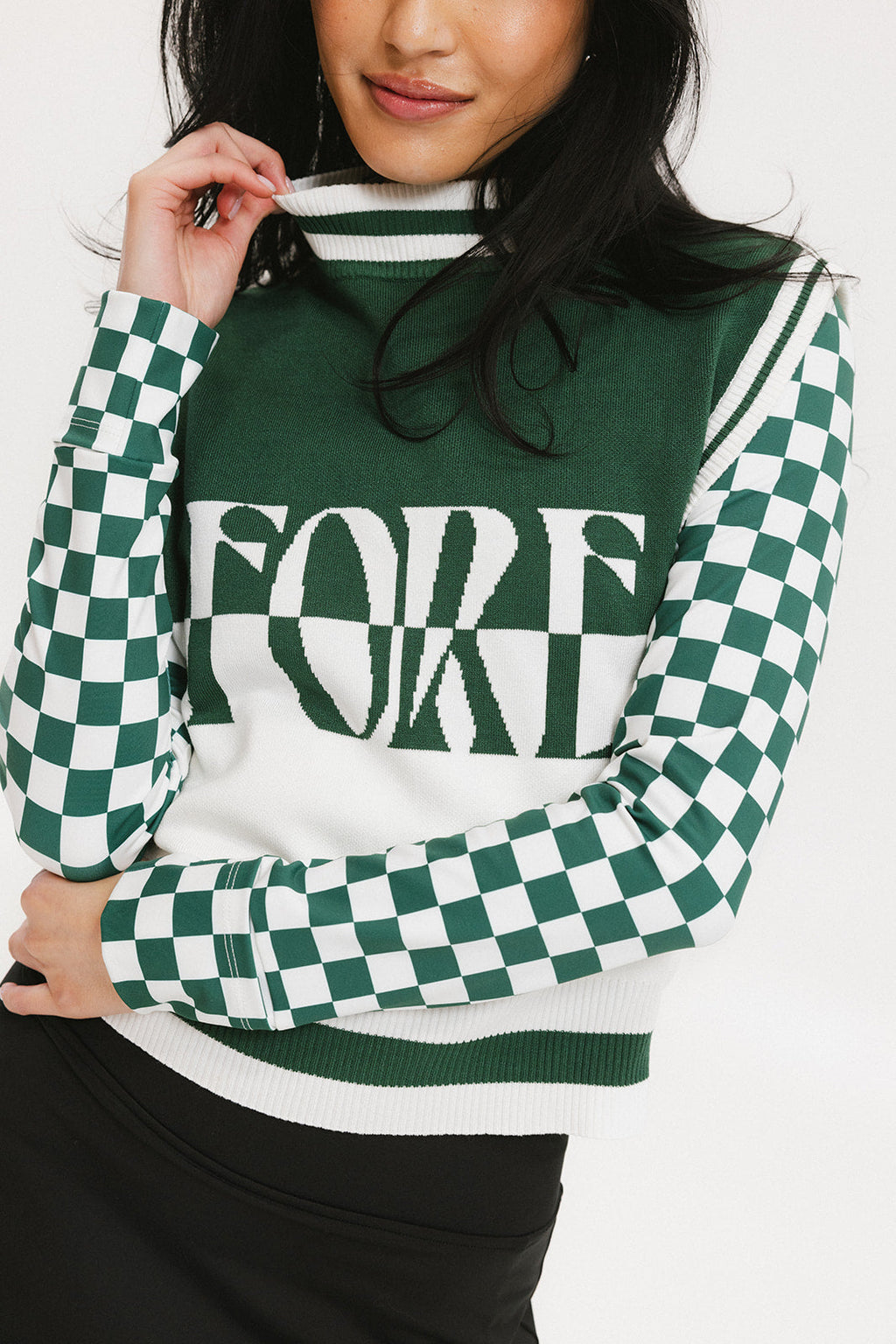 A woman wearing a sleeveless mock-neck sweater with a green and white intarsia knit design and the word 'FORE' written across the front.