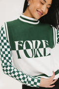 A woman wearing a sleeveless mock-neck sweater with a green and white intarsia knit design and the word 'FORE' written across the front.