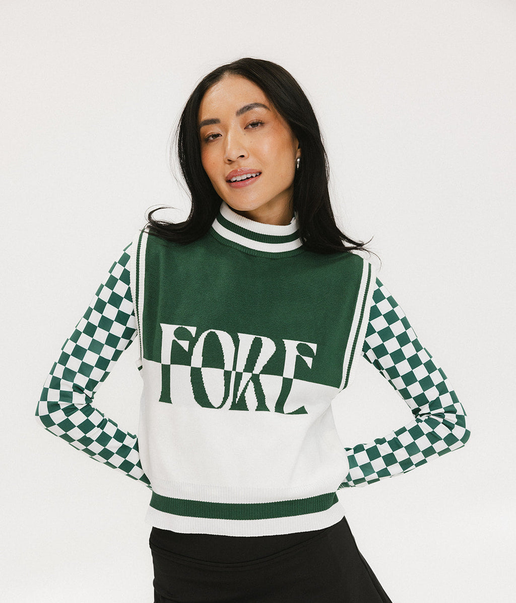 A woman wearing a sleeveless mock-neck sweater with a green and white intarsia knit design and the word 'FORE' written across the front.