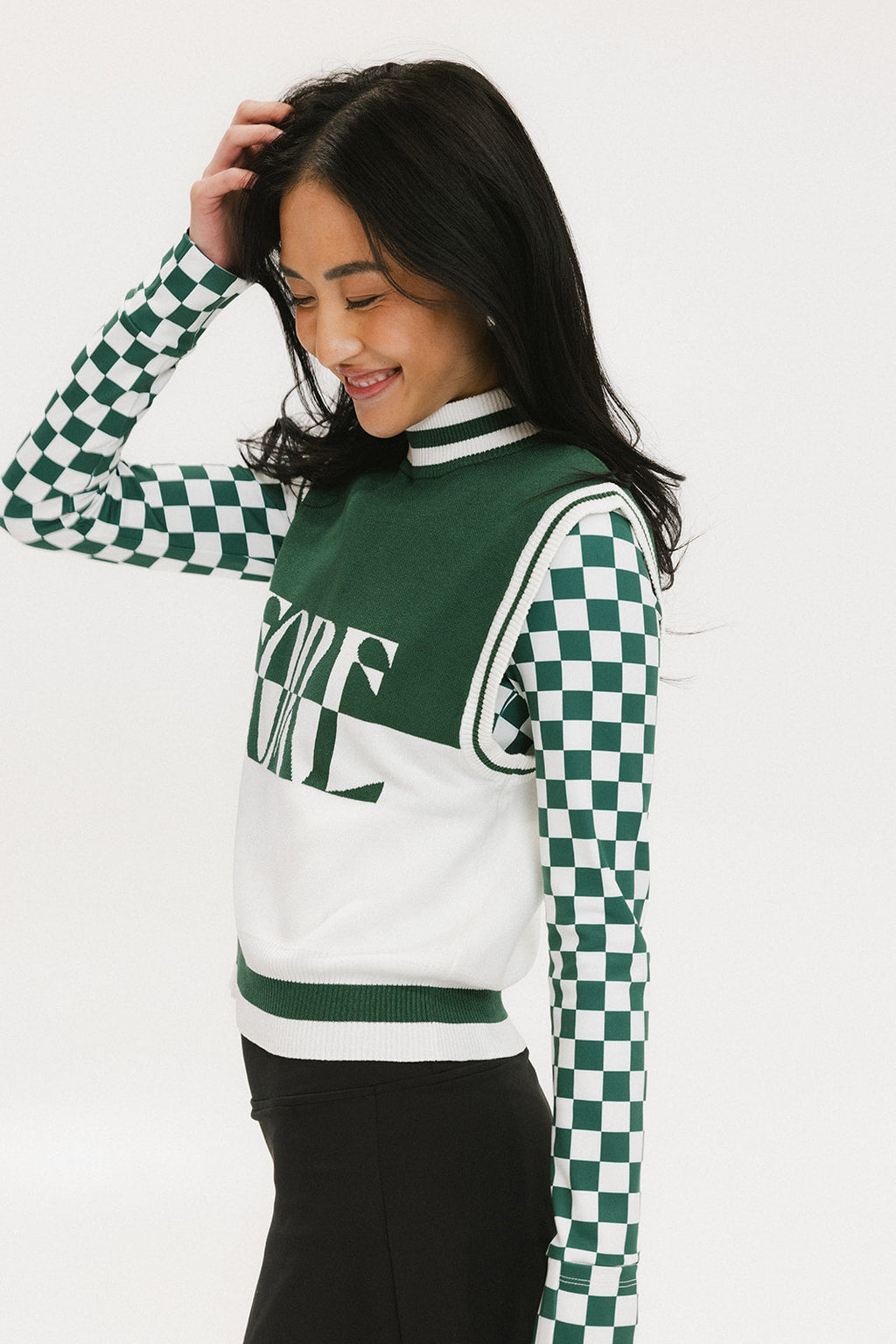 A woman wearing a sleeveless mock-neck sweater with a green and white intarsia knit design and the word 'FORE' written across the front.