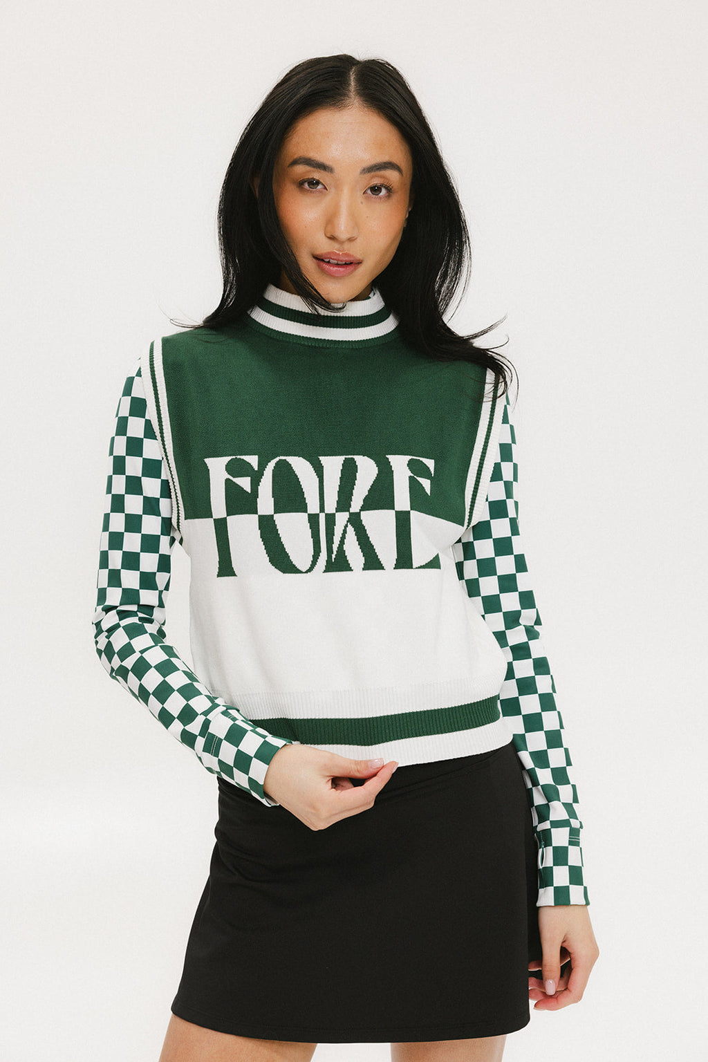 A woman wearing a sleeveless mock-neck sweater with a green and white intarsia knit design and the word 'FORE' written across the front.