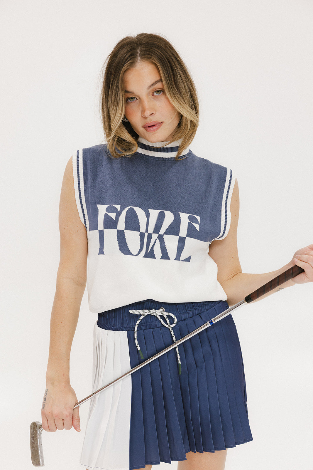 A model wearing an indigo and white intarsia mock neck, sleeveless Fore Sweater.