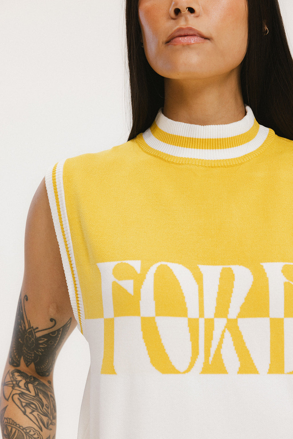 A close-up of a woman wearing a sleeveless sweater with yellow and white colors and the text 'FORE' visible on the front.