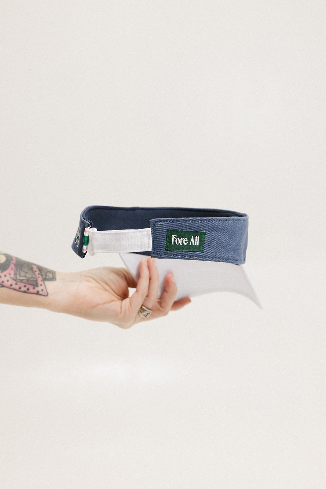 A hand holding a visor with 'FORE' written in bold letters on the front and 'FORE ALL' on the back, against a white background.