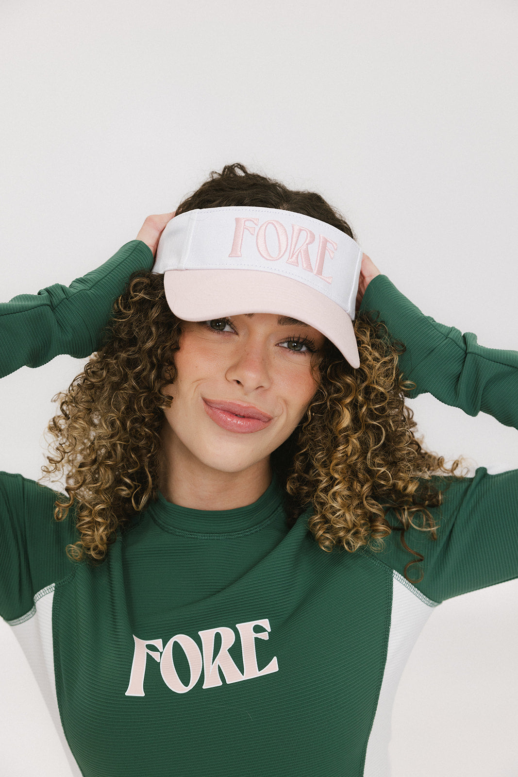 A woman wearing a pink and white 'FORE' visor and a green long-sleeved shirt.