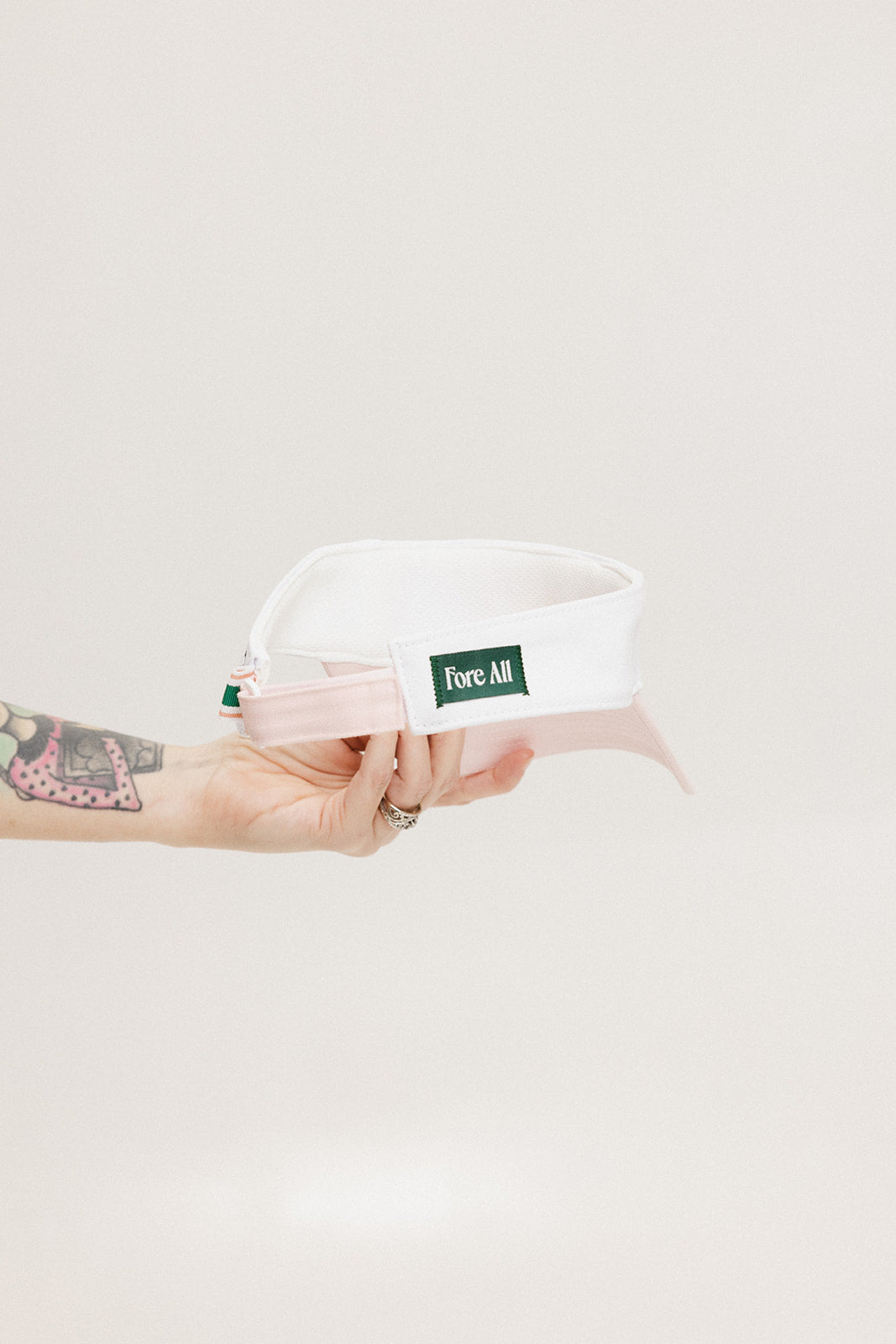 A hand holding a two-toned visor with 'FORE' logo in pink and white colors.