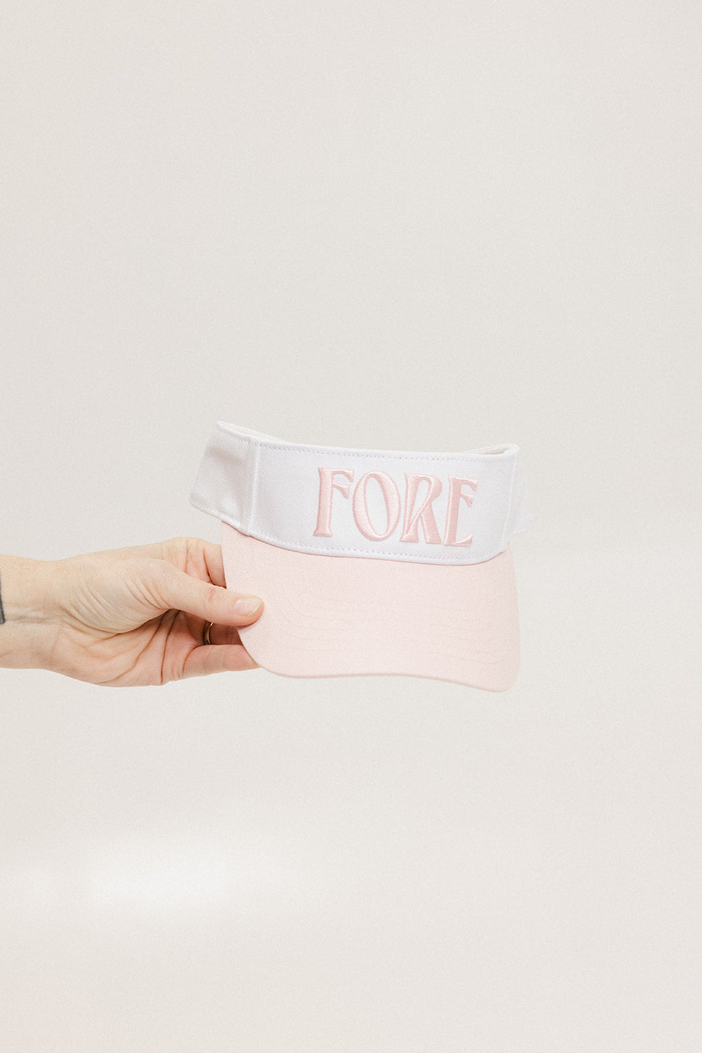 A hand holding a two-toned visor with 'FORE' logo in pink and white colors.