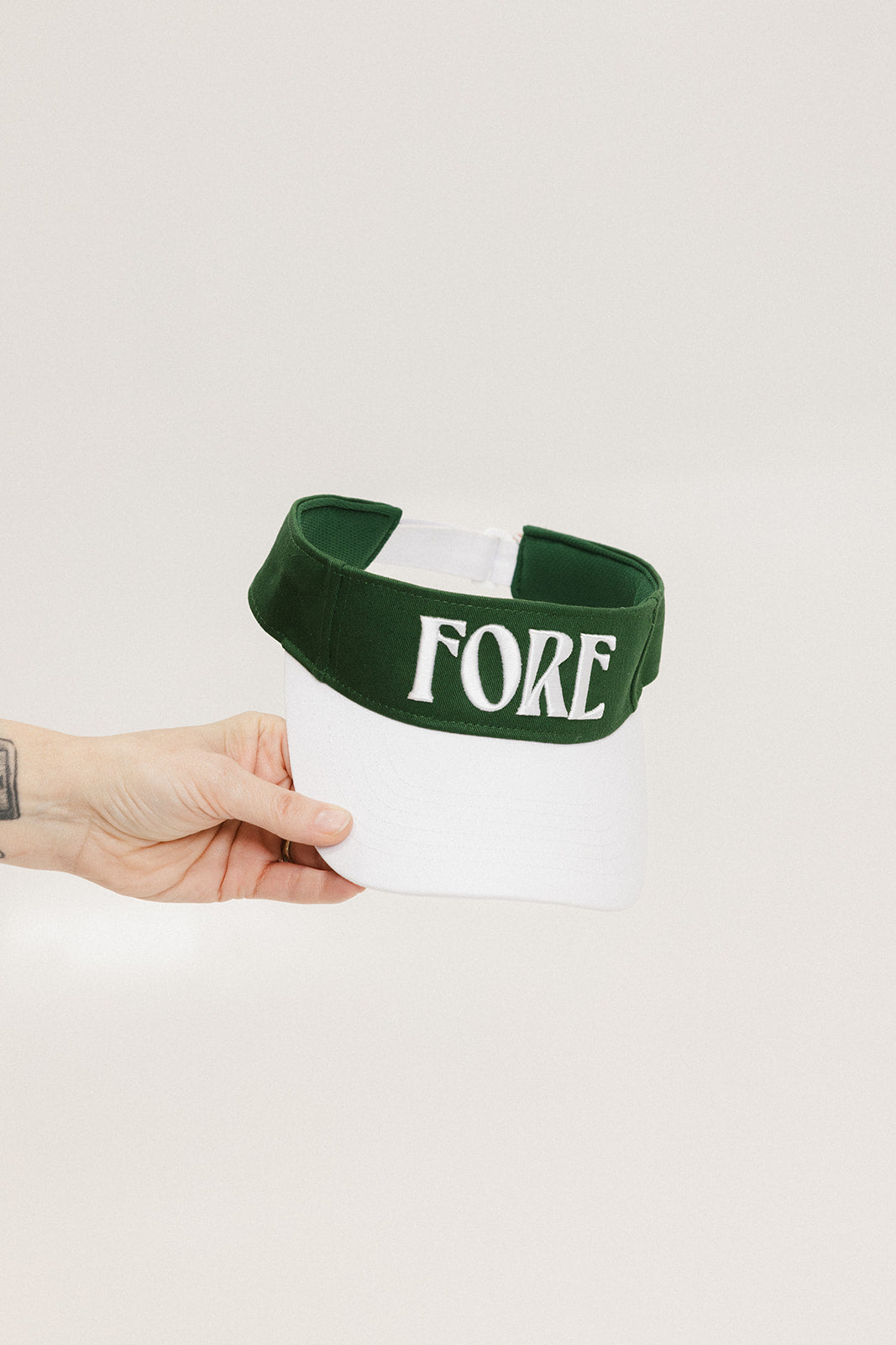 A hand holding a white and green two-toned visor with the word 'FORE' printed on the front.