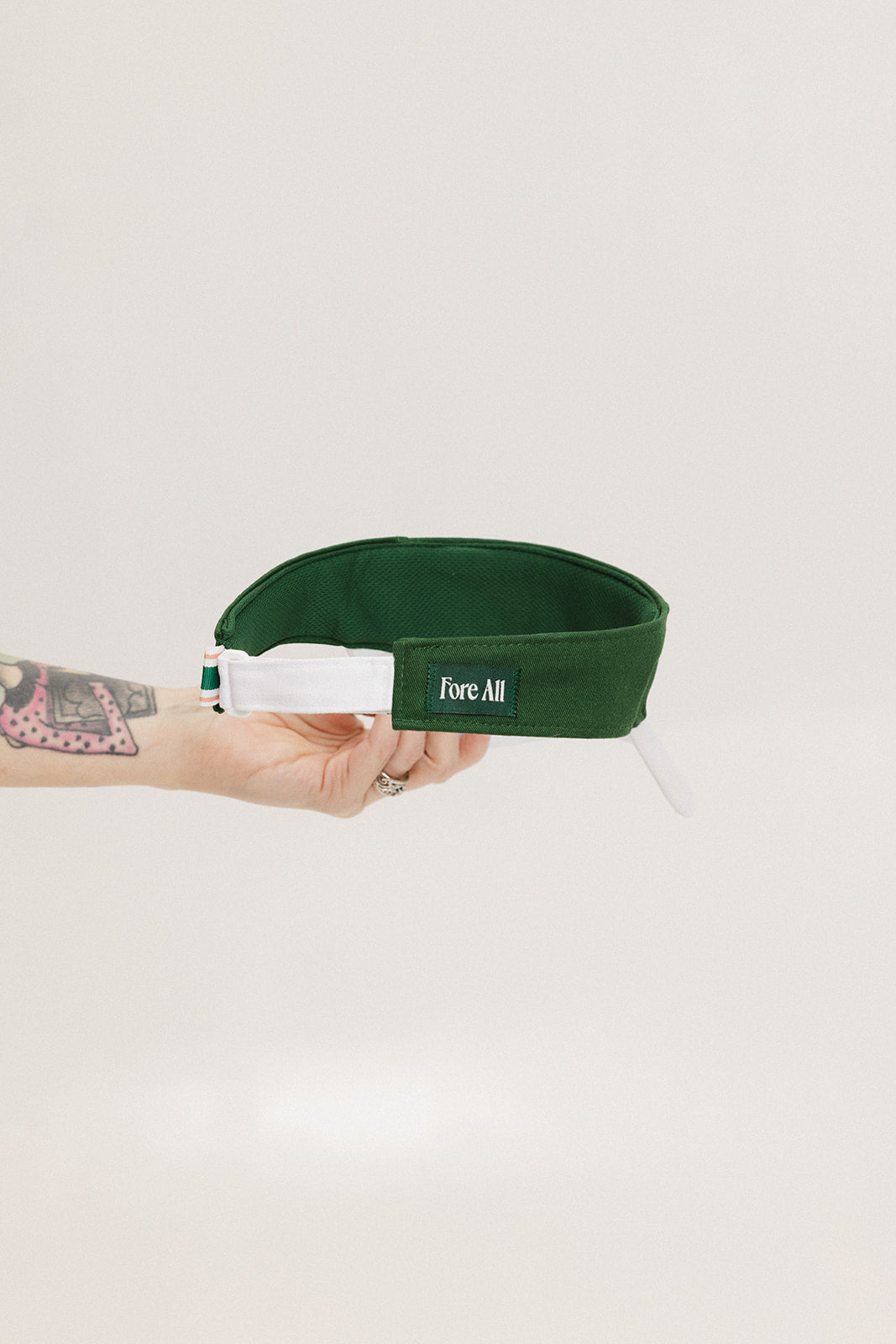 A hand holding a white and green two-toned visor with the word 'FORE ALL' printed on the back.
