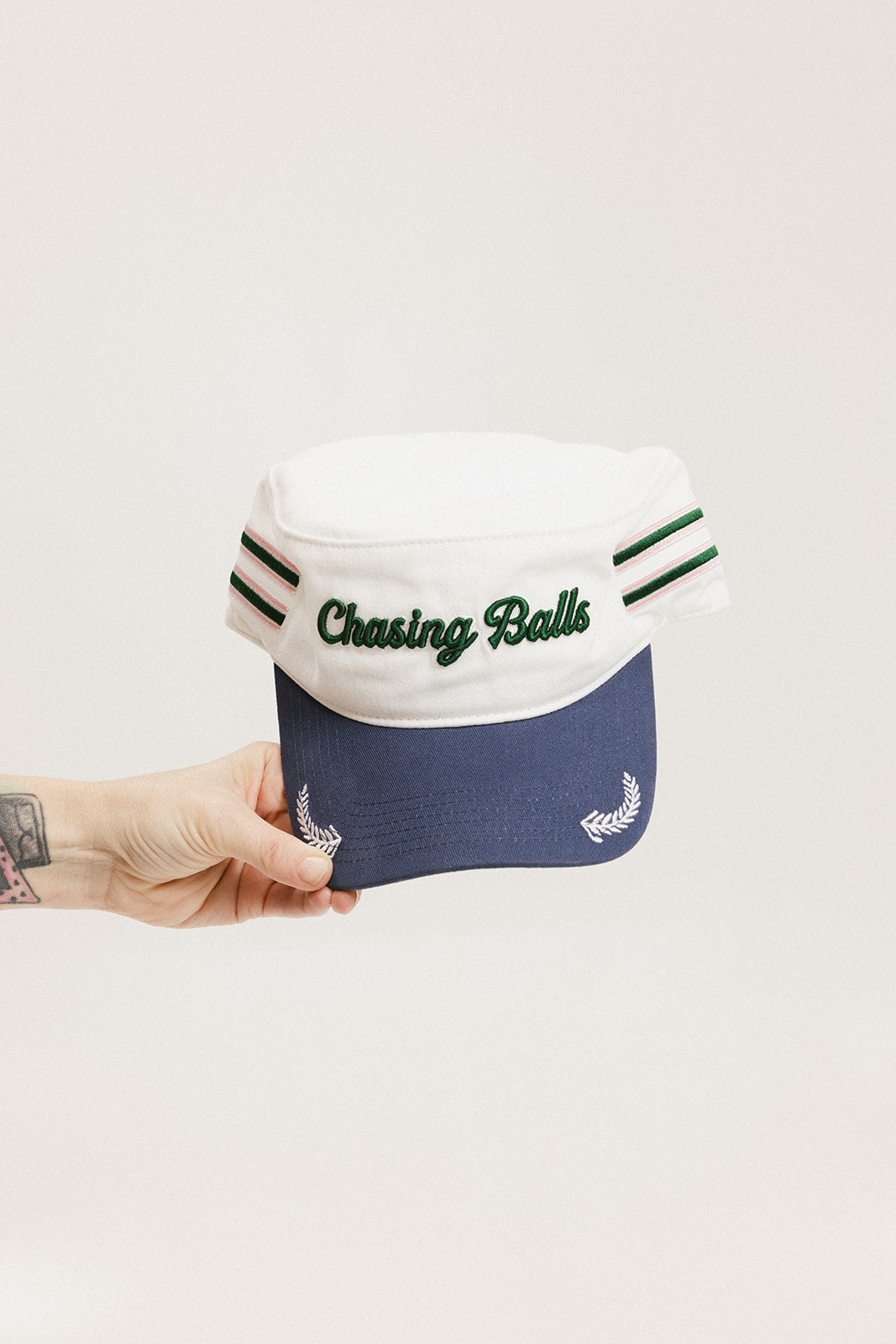 A hand holding a white and blue structured cap with 'Chasing Balls' text and stripes.