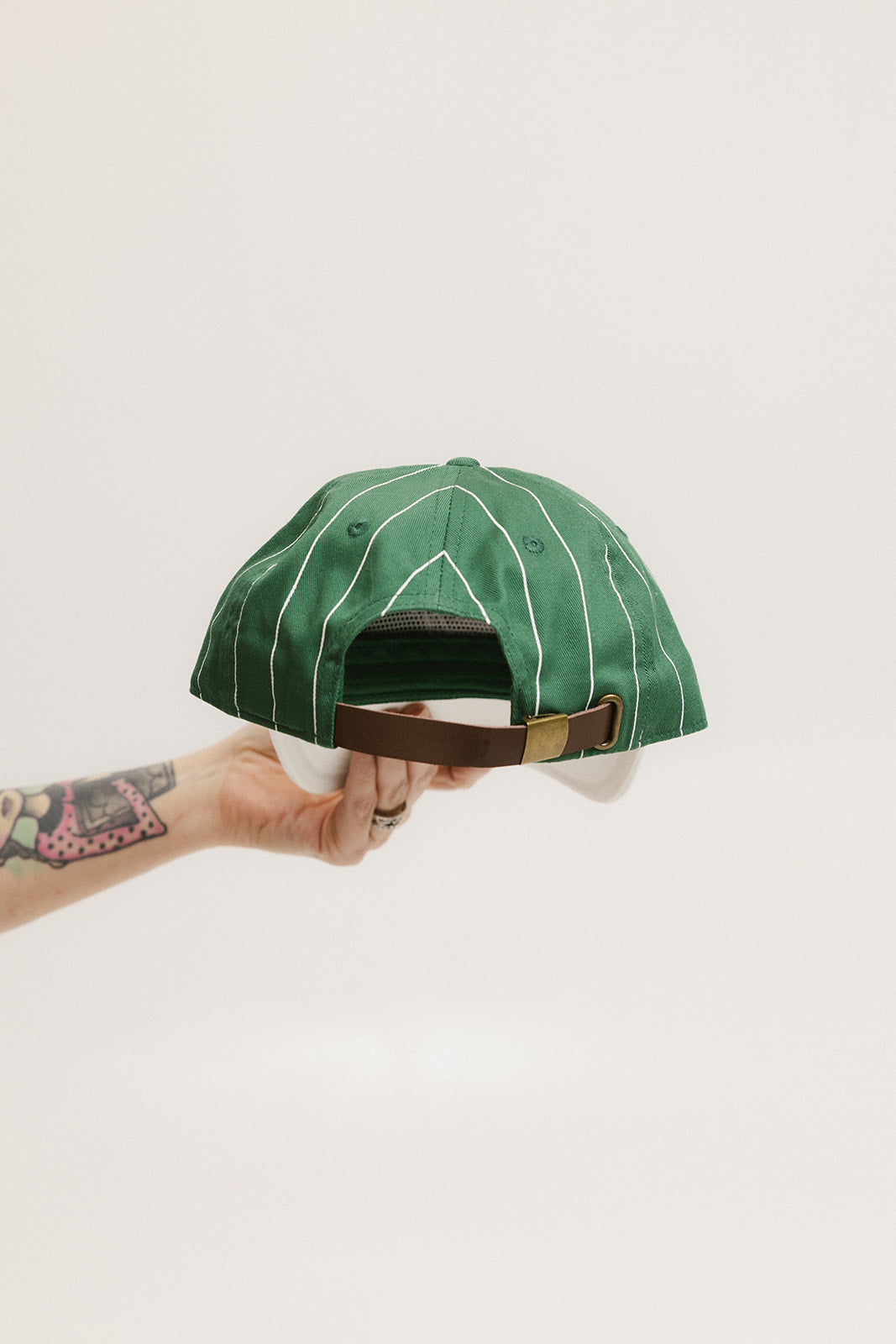 A green baseball cap with pinstripe detailing and an embroidered logo held in a hand against a white background.