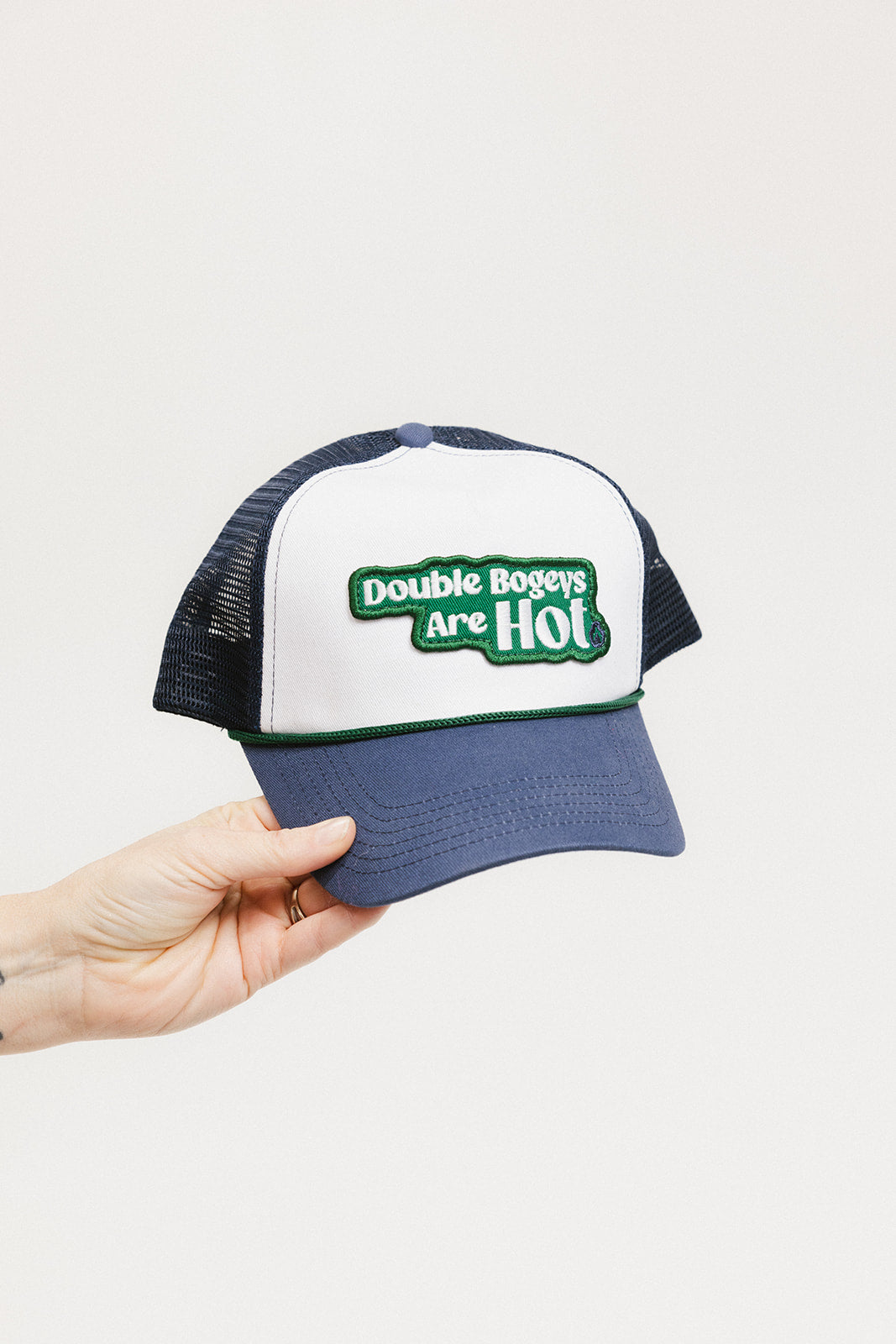 A hand holding a trucker hat with 'Double Bogeys Are Hot' text embroidered on the front, featuring a white panel with green text and a black mesh back.