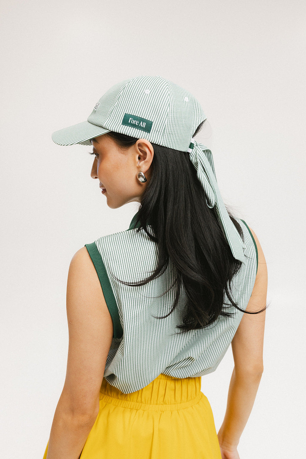 A woman wearing a green seersucker baseball cap with 'Chasing Balls' text embroidered on it and an oversized bow at the back.